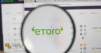 Magnifying glass focusing on the eToro logo on a blurred computer screen, implying analysis or review of the eToro trading platform.