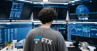 Sam Bankman-Fried, wearing an FTX t-shirt, monitors multiple trading screens displaying cryptocurrency market data.