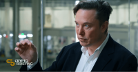 Elon Musk speaking and gesturing with his hand in an industrial setting.