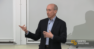 Gary Gensler speaking in a classroom setting, gesturing with his hands in front of a whiteboard.